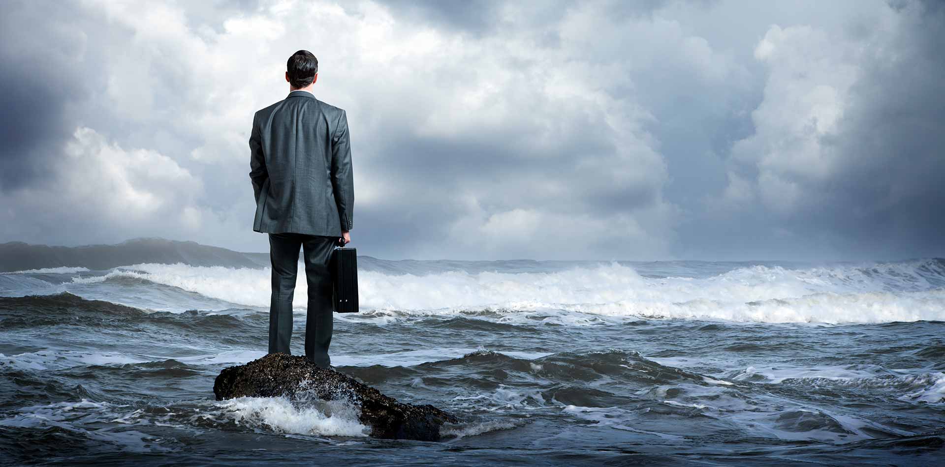 A man in a suit holding a briefcase. He stands on top of a rock in the ocean while looking out into the distance.