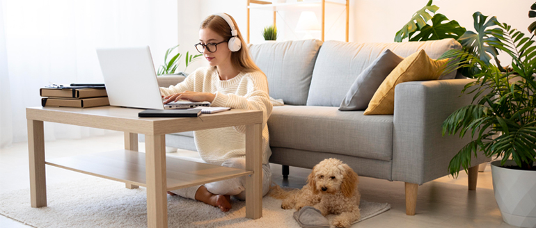 A young woman sitting on her rug while working on her laptop with her headphones in. Her dog is laying beside her.