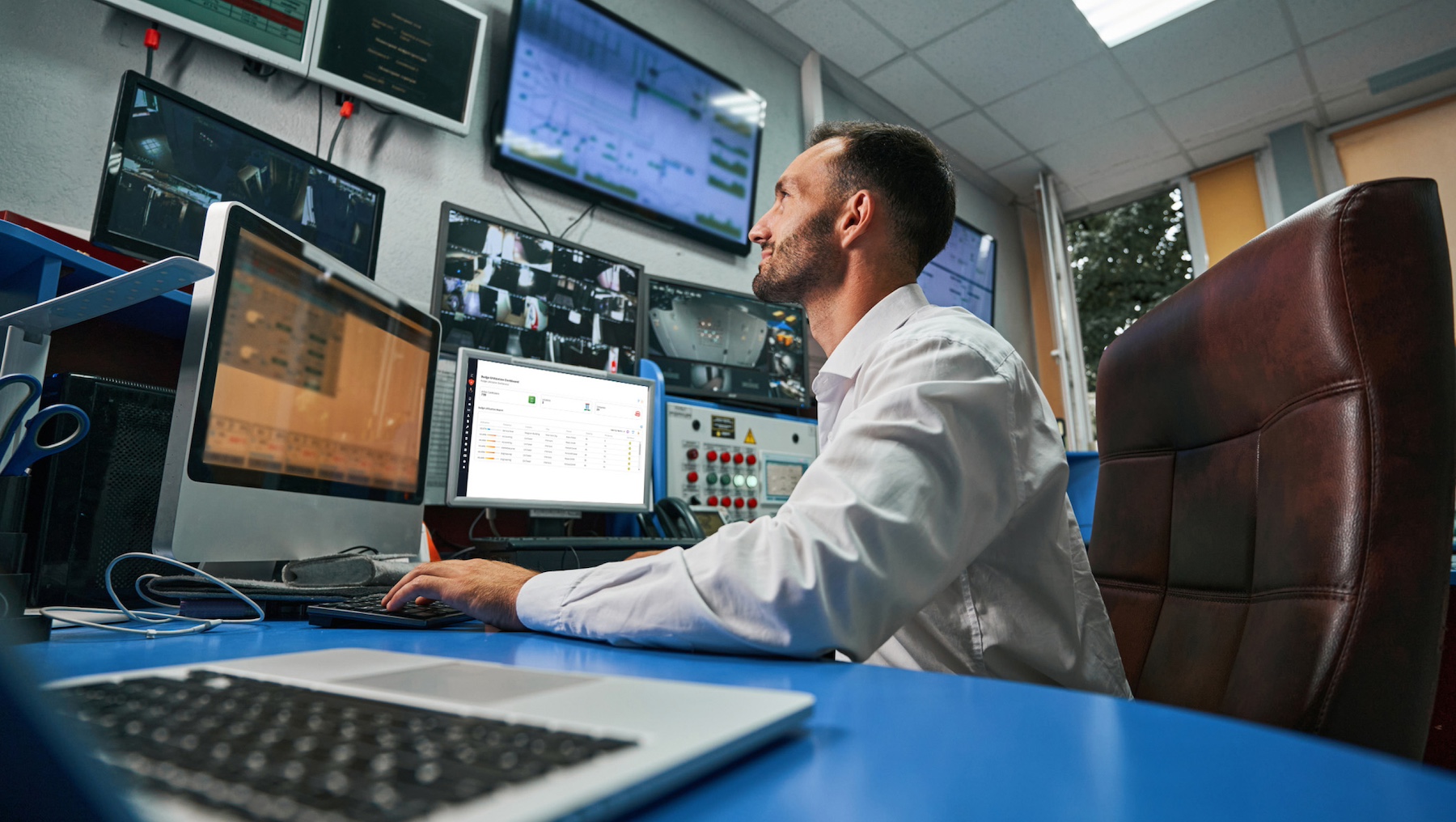 A person in a control room monitors multiple screens displaying data and video feeds, seated at a desk with a laptop and various equipment.