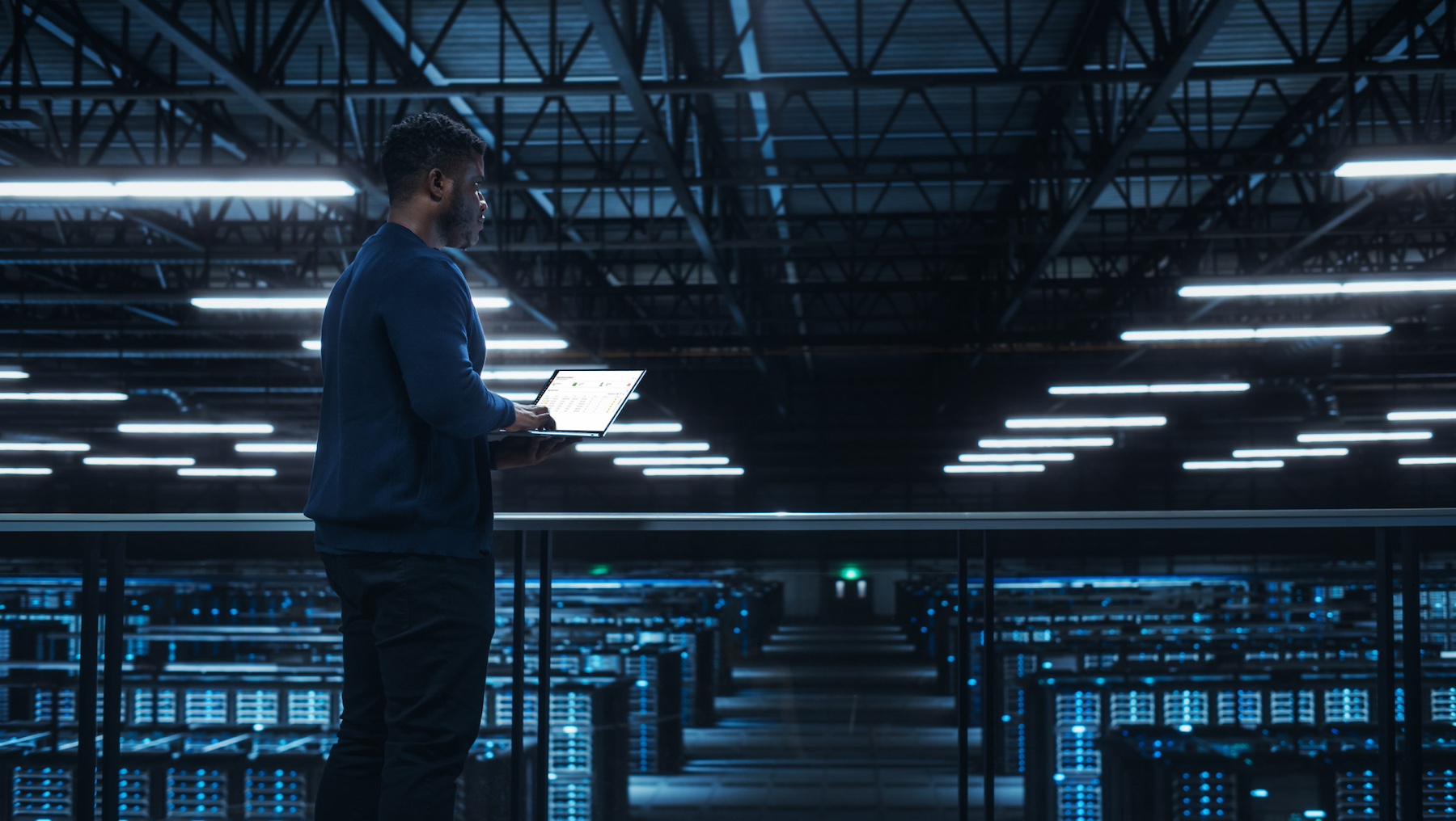 A person stands in a dimly lit data center holding a laptop, overlooking rows of server racks with illuminated blue lights.