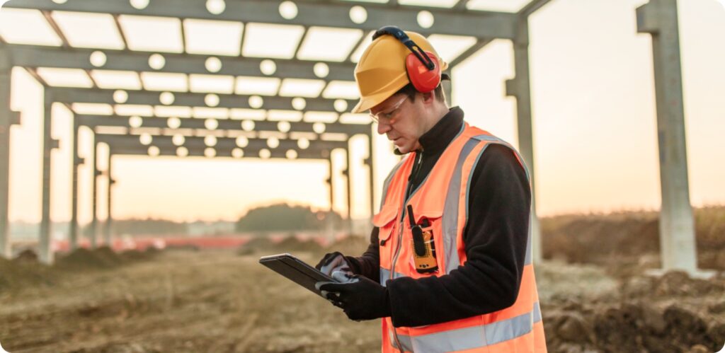 A man in an orange vest and hard hat inputting information into his IPad at a construction site.
