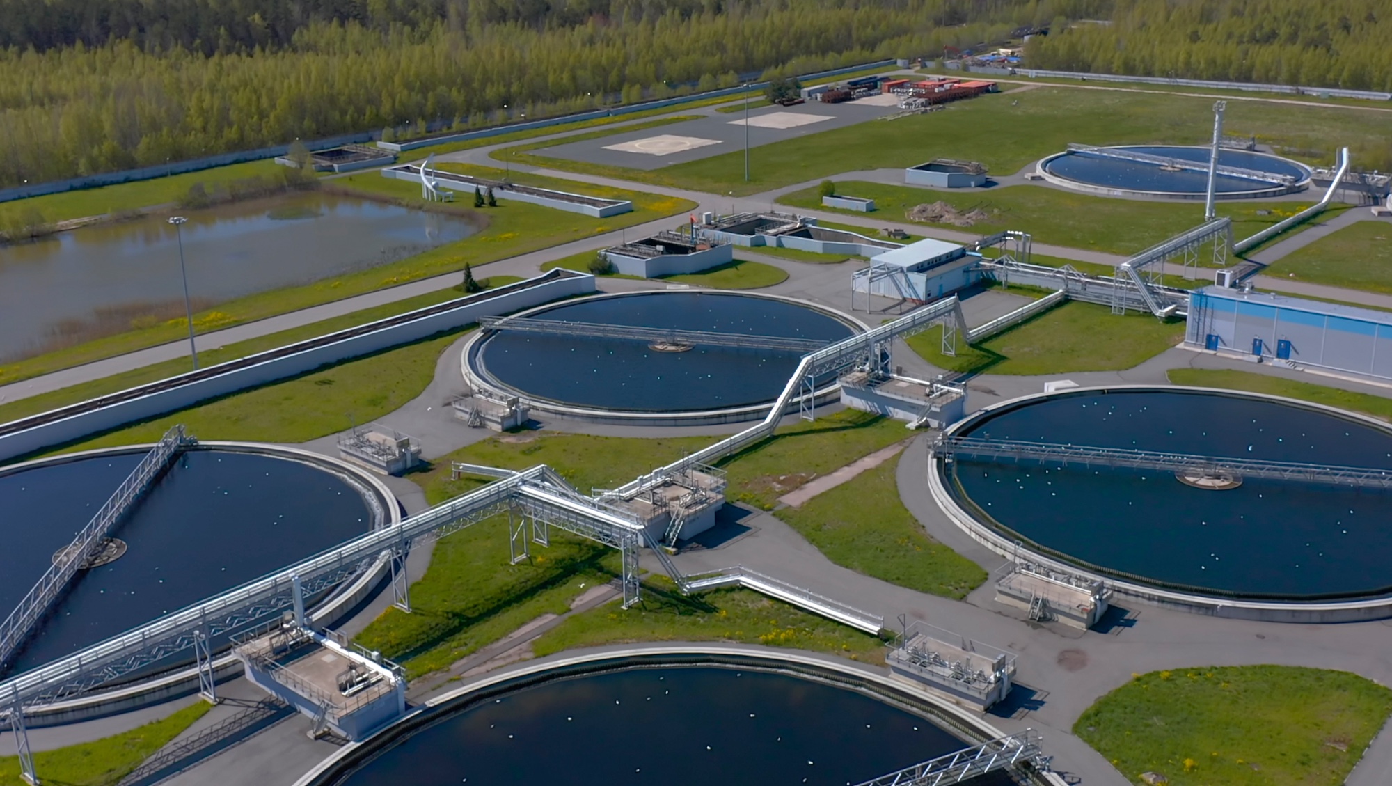 Aerial view of a wastewater treatment plant with large circular tanks and pipelines, surrounded by green fields.