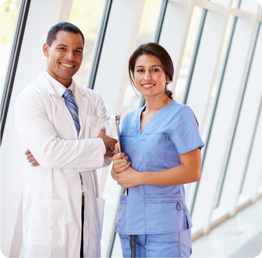 A doctor in a white coat and a nurse in blue scrubs stand side by side in a brightly lit hallway.