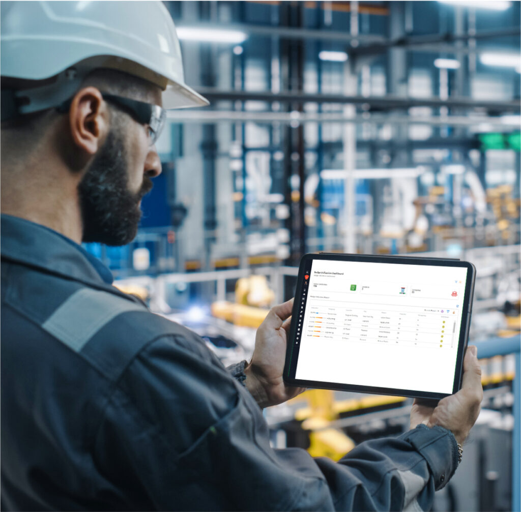 A worker in a hard hat views a digital tablet displaying data in an industrial setting with machinery in the background.