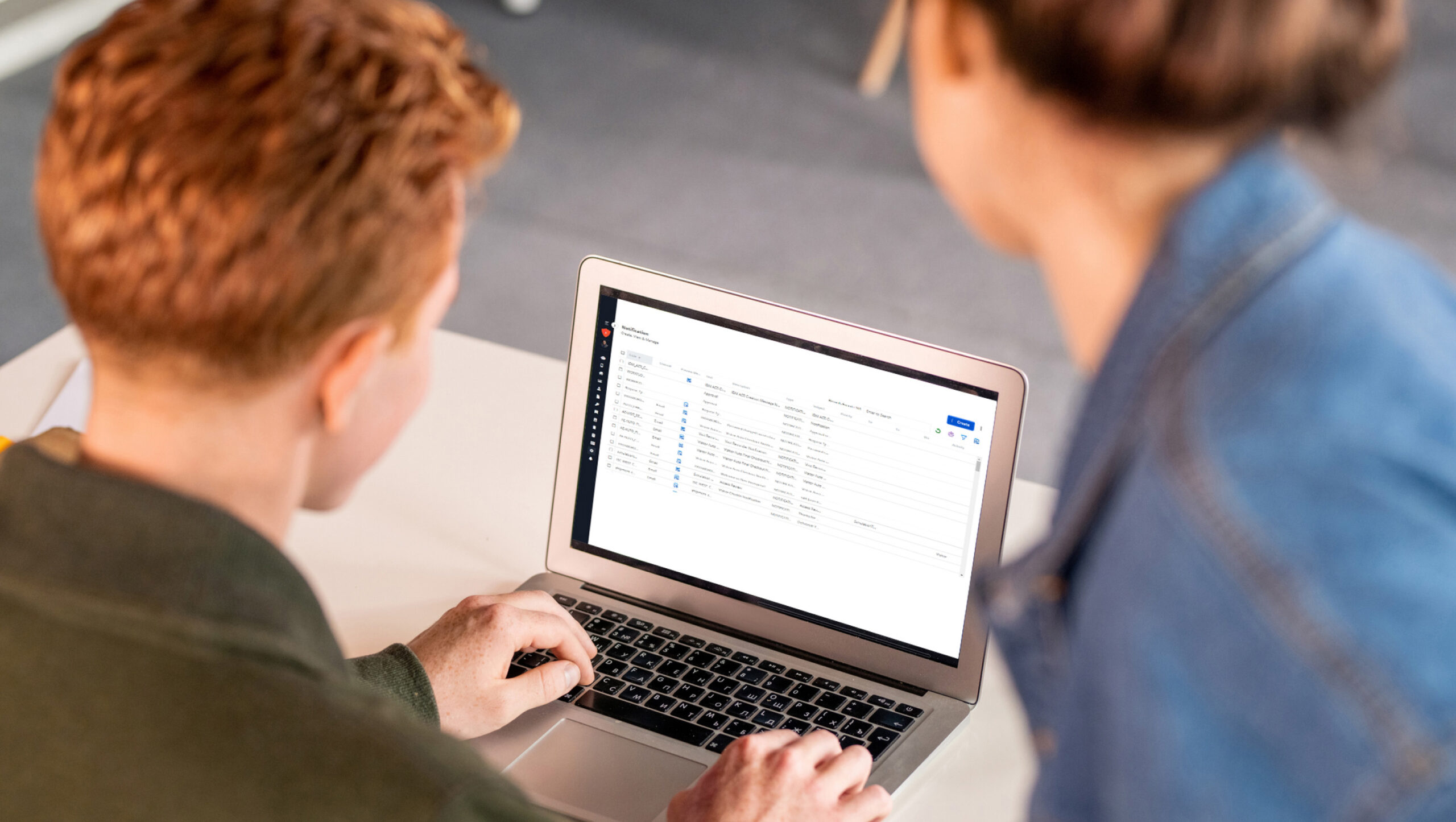 Two people using a laptop, one typing while the screen displays a spreadsheet or database.