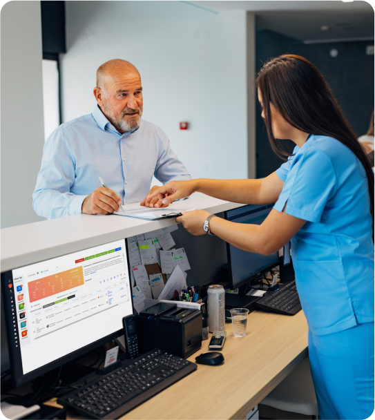 A man in a blue shirt signs a document handed by a woman in scrubs at a reception desk equipped with computer monitors, ensuring compliant visitor management.