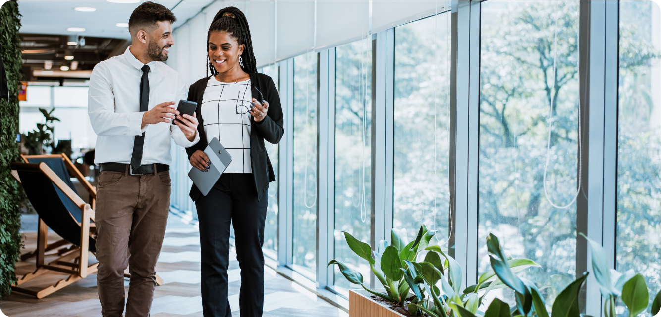 Two people walking in an office hallway, holding a tablet and smiling, with large windows and plants in the background.