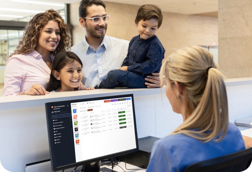 A family stands at a reception desk smiling at a receptionist. A computer monitor displays a software dashboard.