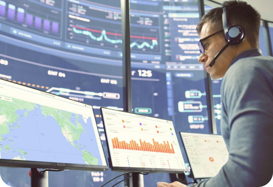 A person wearing a headset is focused on multiple computer screens displaying graphs and data in a control room setting.