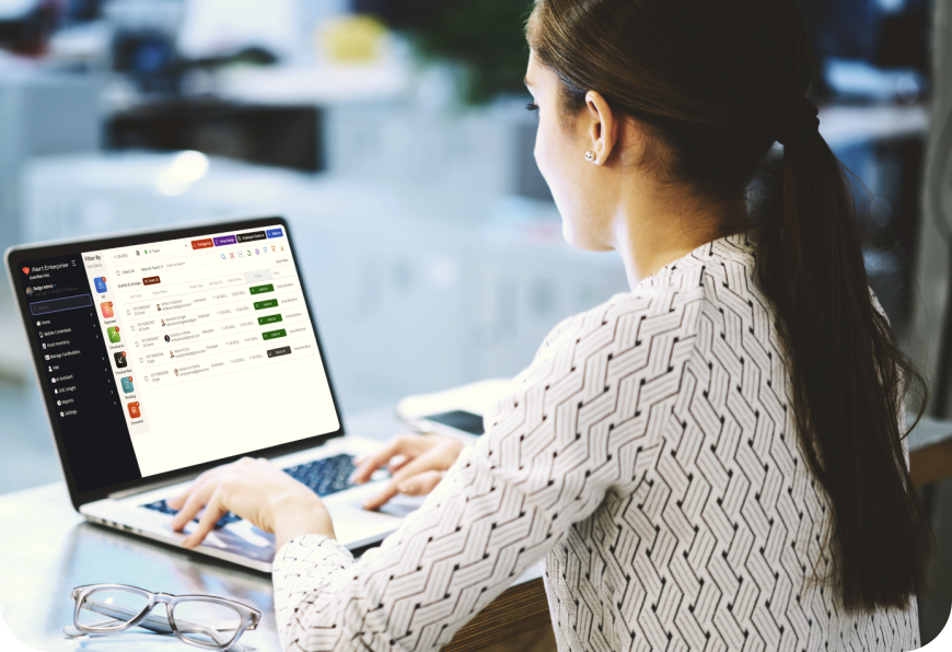 A woman in a patterned blouse is working on a laptop at a desk. The laptop screen displays a task management software interface. Glasses are placed on the desk beside her.