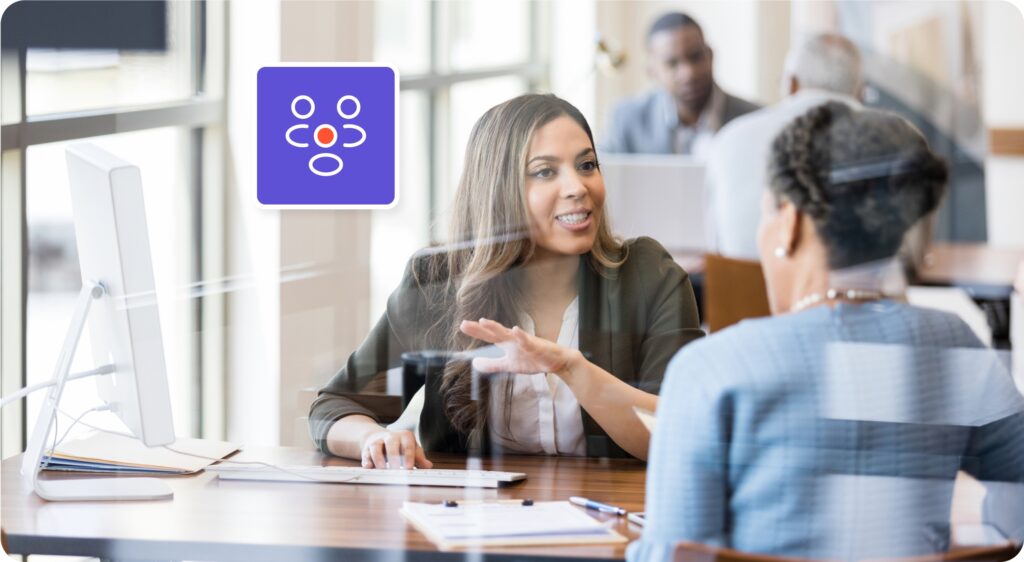 Two people are sitting at a desk in an office setting, having a conversation. There is a computer monitor and a framed icon on the desk.