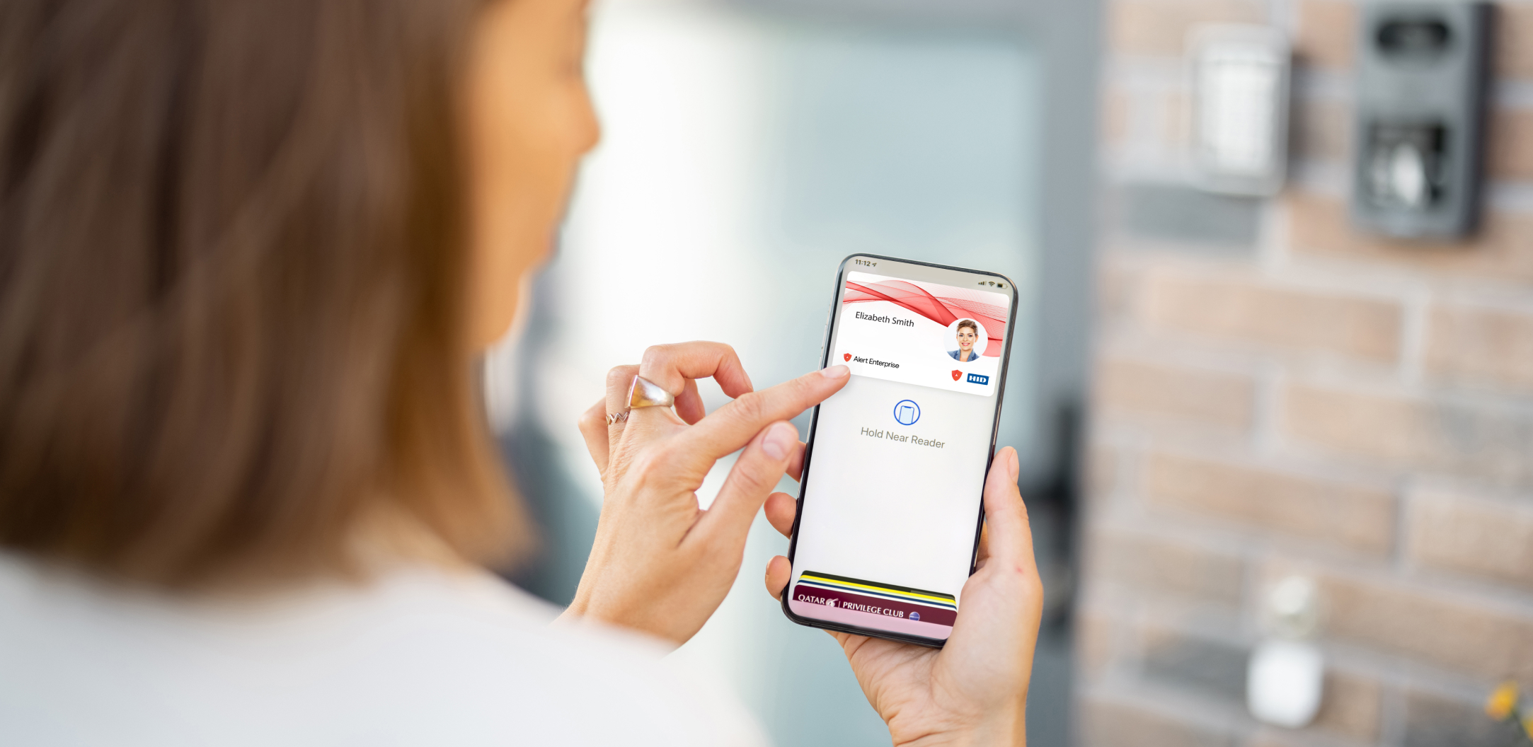 A person holds a smartphone displaying a digital ID on the screen, positioned near a card reader on a brick wall.