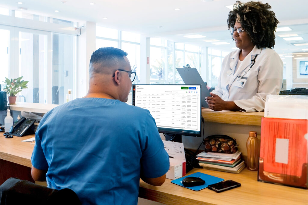 A healthcare professional in a white coat stands at a desk, holding a clipboard, while another sits at a computer, viewing a medical schedule in a bright, modern office.