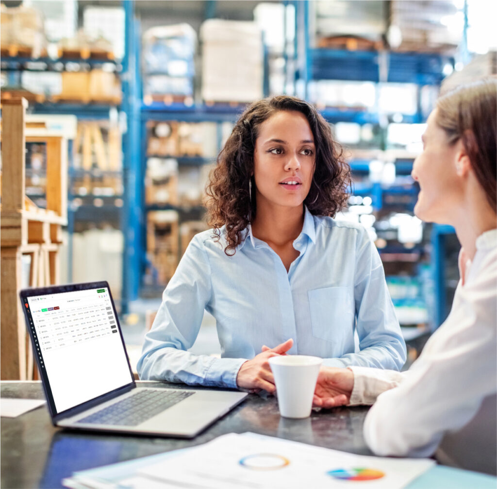 Two women discussing at a table in a warehouse setting, with a laptop displaying a spreadsheet and papers nearby.