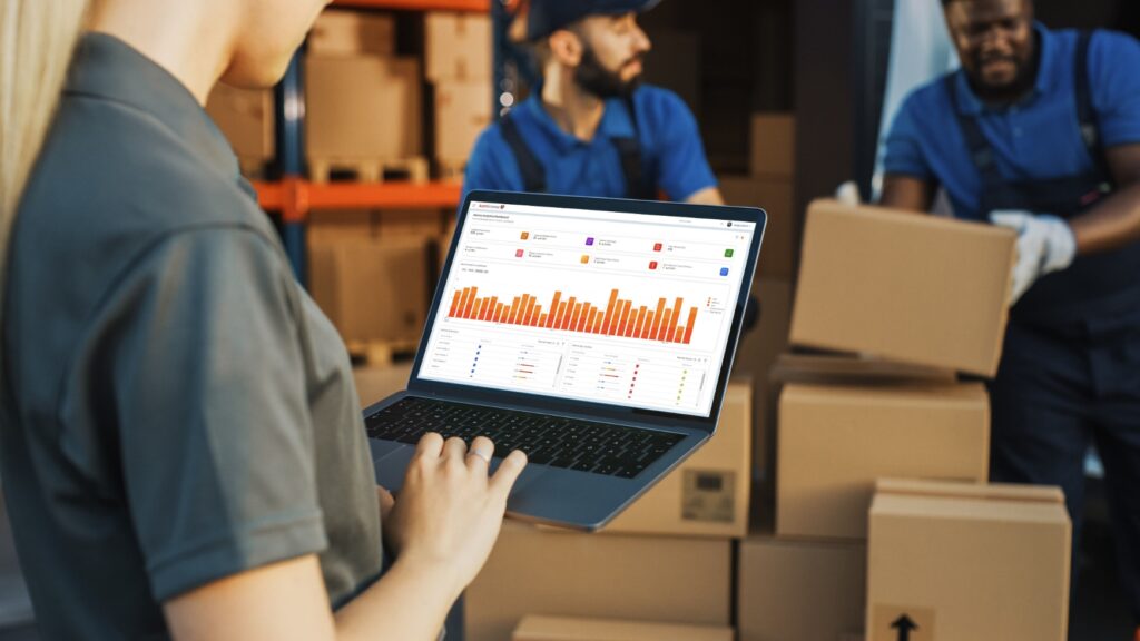 A person in a warehouse holds a laptop displaying a bar graph. In the background, two workers move cardboard boxes.