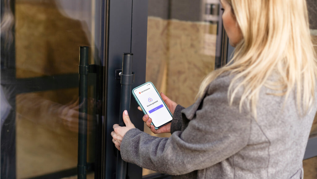 A person with blonde hair uses a smartphone to unlock a glass door.