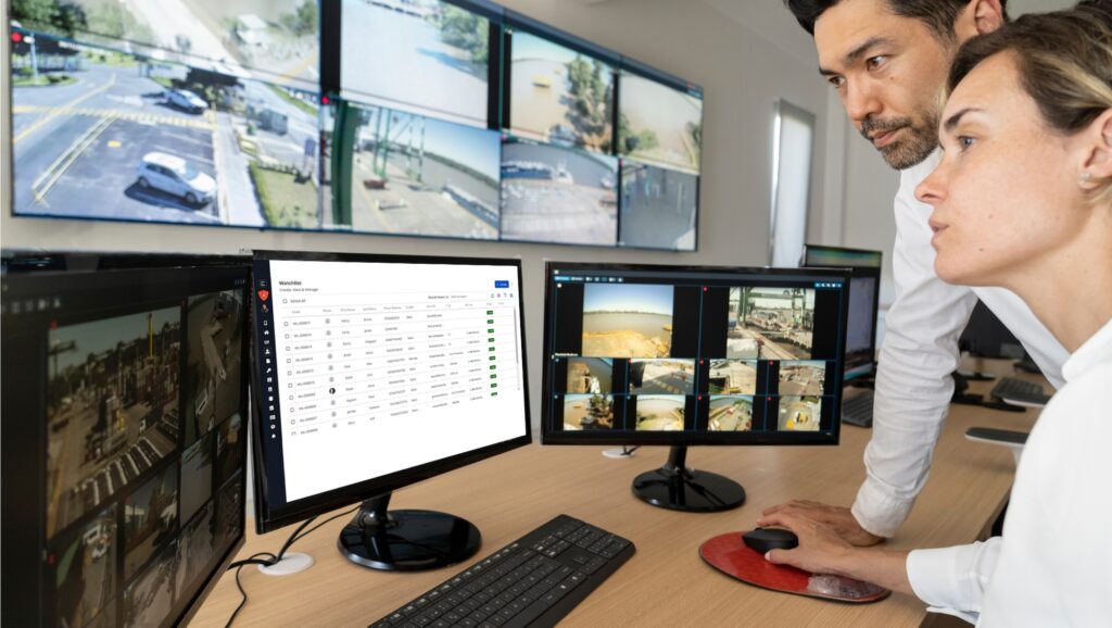 Two people monitor multiple surveillance screens in a control room.