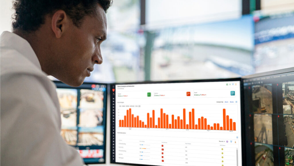 A person analyzes data on computer screens displaying graphs and security footage in a control room.