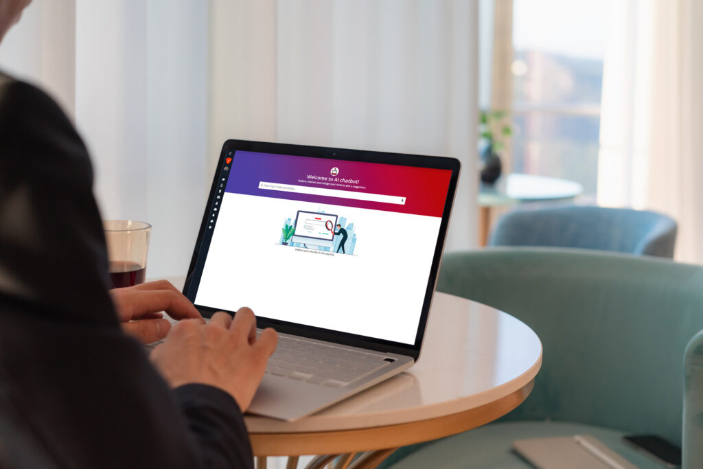 Person using a laptop displaying a white screen with a red and purple header and a graphic of two people beside a large computer screen on a round table.