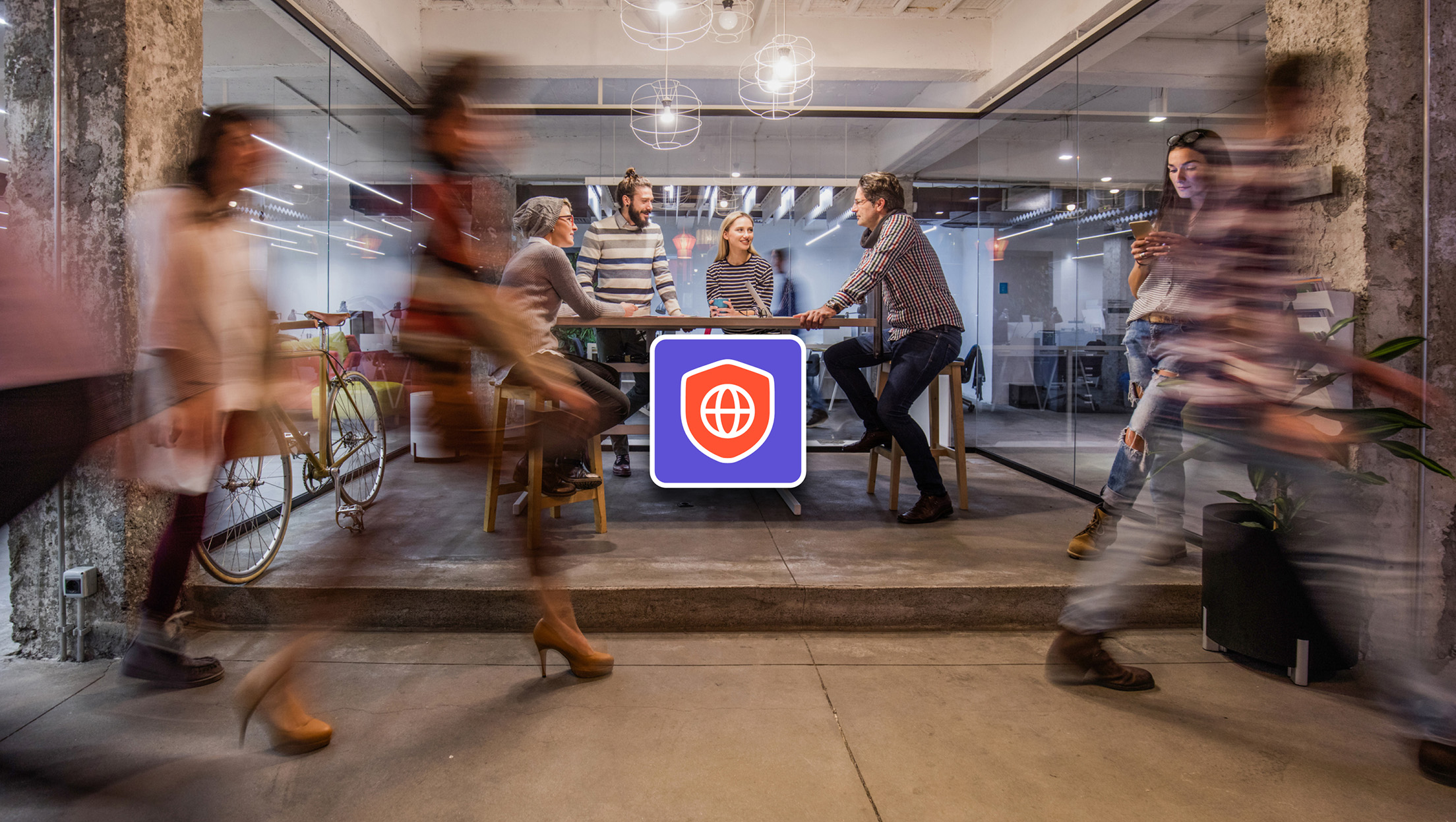 A group of people is having a meeting at a table in a modern office space. Several people walk by in the foreground.