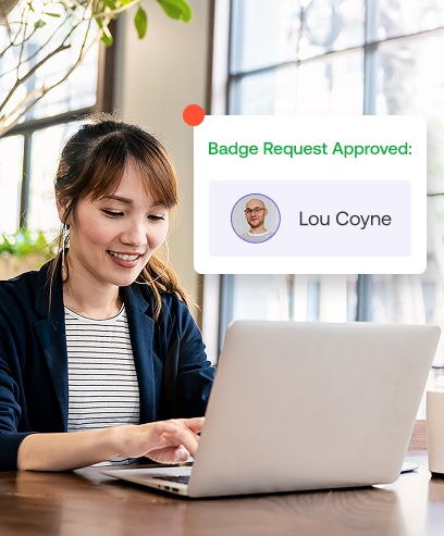 A woman is smiling while using a laptop. An on-screen notification shows a badge request approved for Lou Coyne.