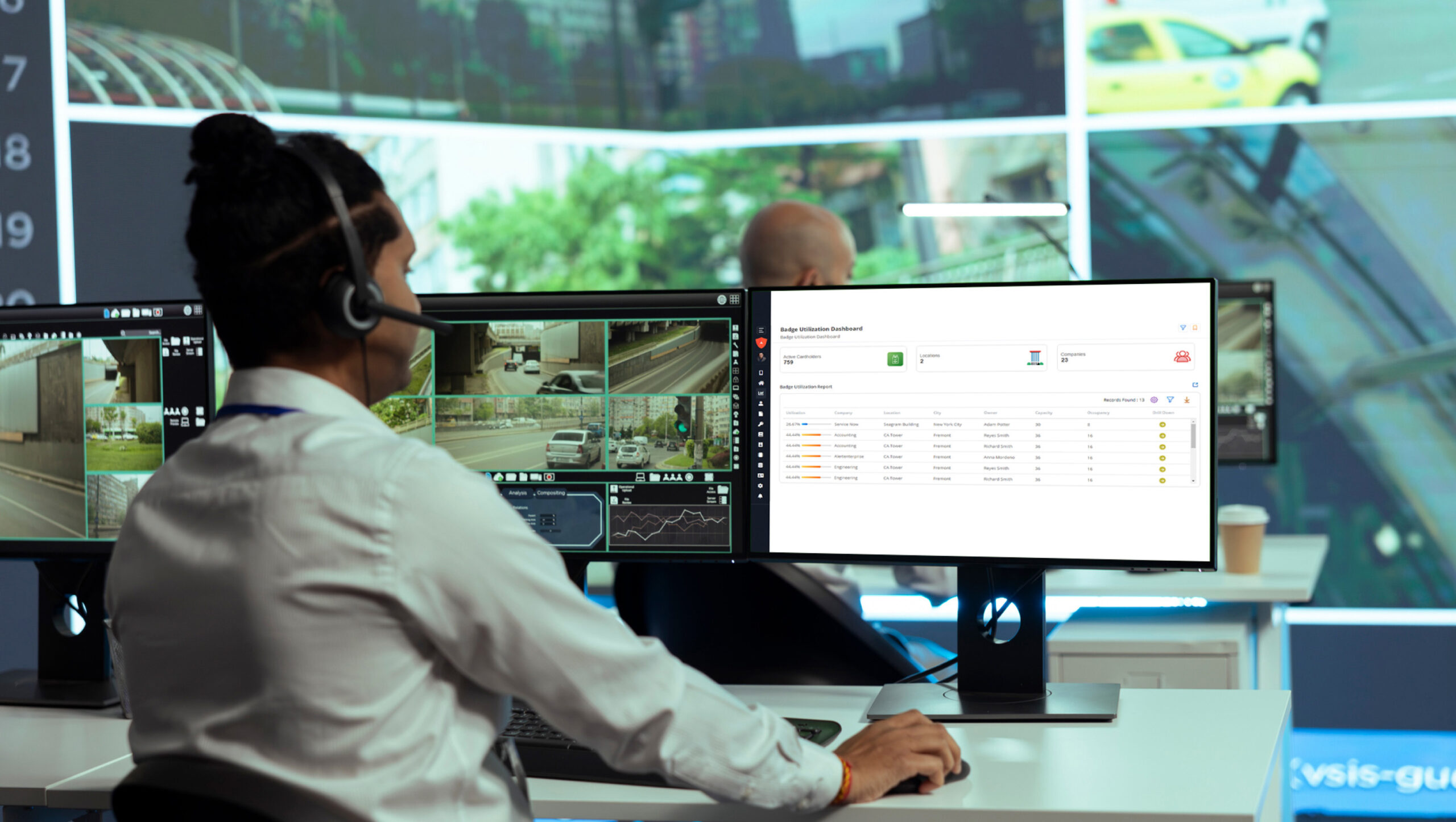 A person with a headset monitors multiple surveillance screens and a digital dashboard in a control room.