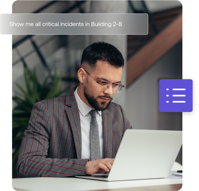 A man in a gray suit and glasses is working on a laptop in an office setting. A digital overlay displays a query for critical incidents in Building 2-B.