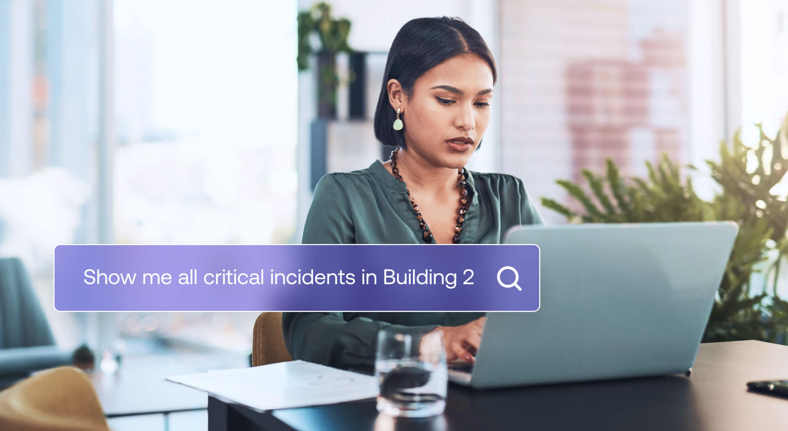 A woman sits at a desk working on a laptop. The screen displays a search bar with the text, Show me all critical incidents in Building 2.