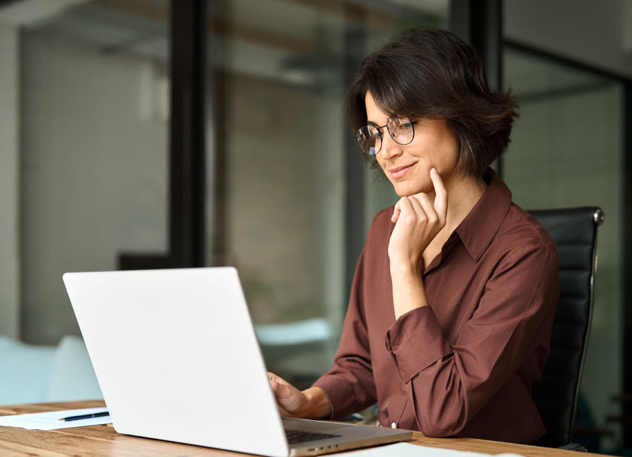 Person seated at a desk, looking at a laptop with a thoughtful expression. They are wearing glasses and a brown shirt, in a modern office setting.