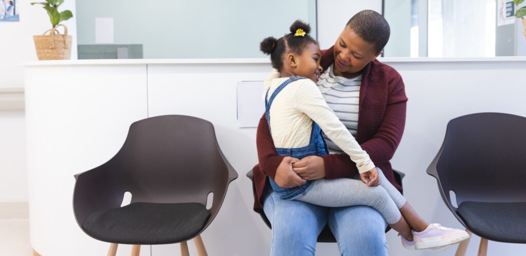 An adult and a child sit together in a waiting area, with the child on the adult’s lap. They appear to be smiling and interacting affectionately. Two empty chairs are nearby.