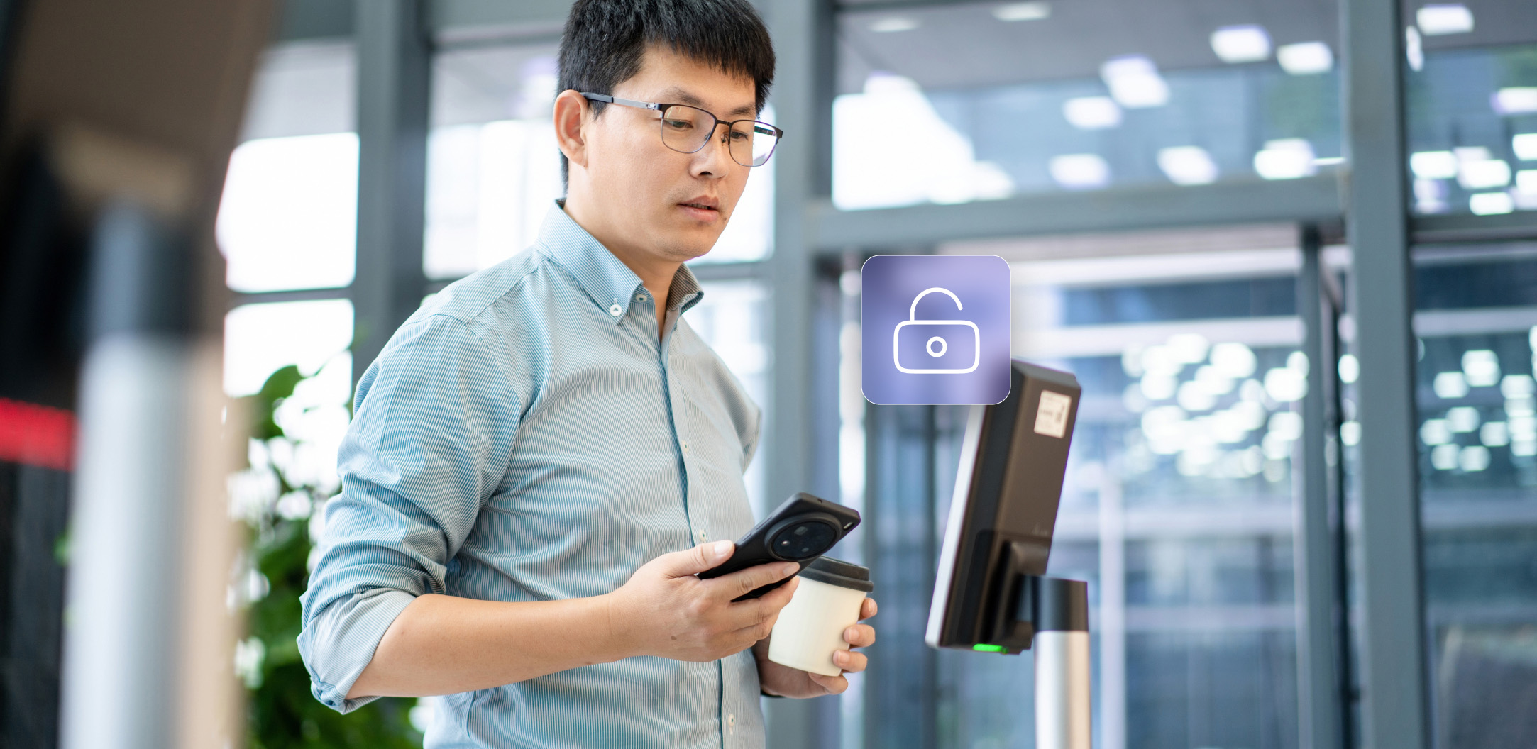 A man holding a smartphone and a coffee cup stands in front of a digital kiosk, with an unlocked padlock icon displayed between him and the screen.