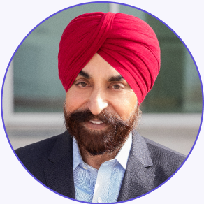 A man with a red turban, beard, and mustache is wearing a dark suit and light blue shirt, standing in front of a blurred background.