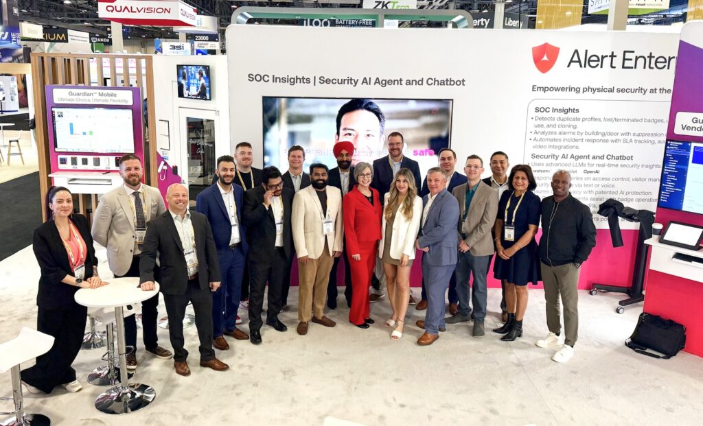 A group of people in business attire pose for a photo in front of a trade show booth with “Alert Enterprise” and security technology displays.