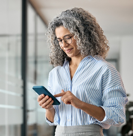 A woman with curly gray hair and glasses is standing indoors, looking at and using a smartphone. She is wearing a blue and white striped shirt.