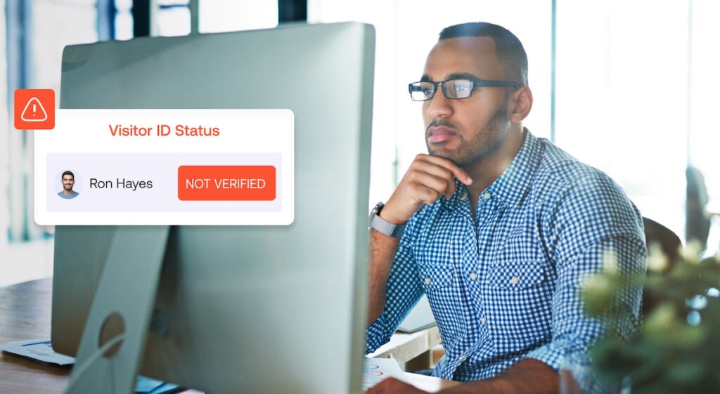 A man wearing glasses works at a computer. Onscreen, a pop-up indicates Visitor ID Status: Ron Hayes, NOT VERIFIED with a warning icon.