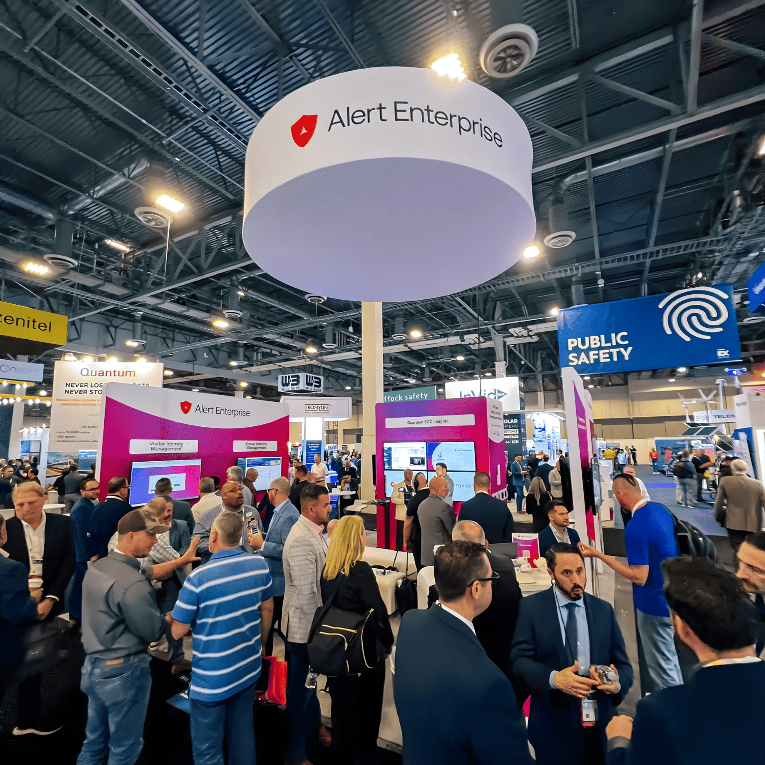 Crowd of people networking and visiting the Alert Enterprise booth at a busy trade show, with various company displays visible in the background.