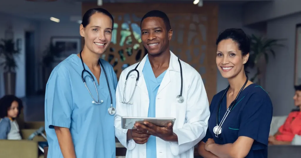 Three healthcare professionals, two women and one man, stand together indoors, wearing scrubs and lab coats with stethoscopes, smiling at the camera.