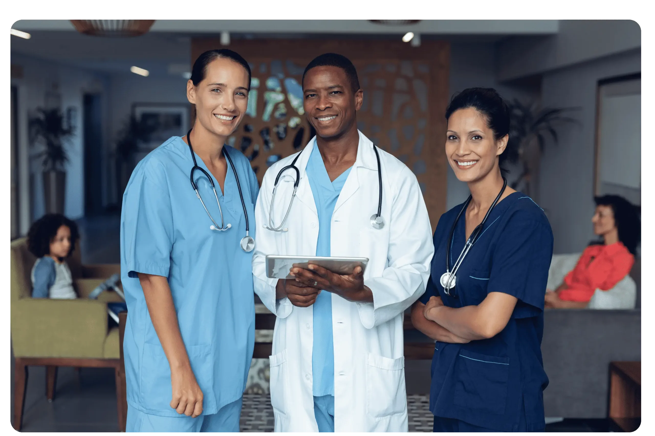 Three medical professionals, two women and one man, stand together indoors wearing scrubs and lab coats, smiling and holding a tablet, with people seated in the background.