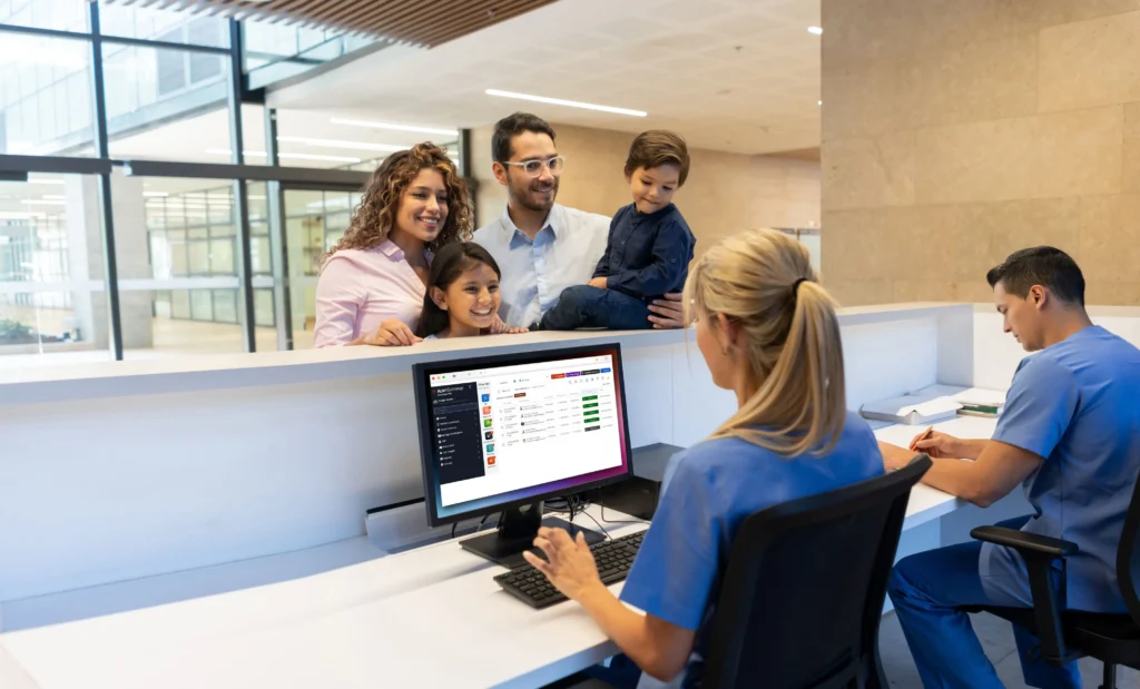 A family stands at a medical reception desk while a staff member in scrubs uses a computer and another writes in a notebook.