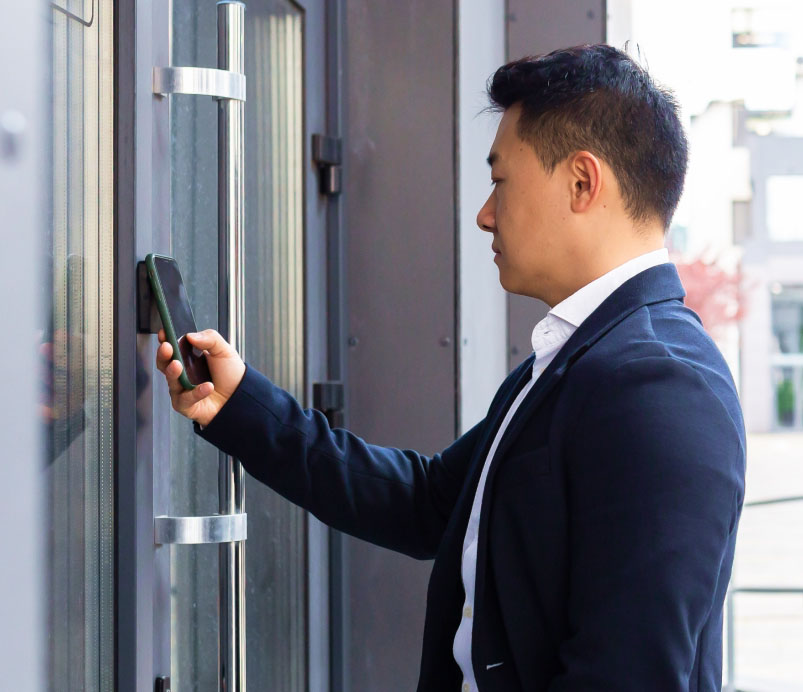 A man in a suit uses a smartphone to unlock a door by holding it up to an electronic access panel.