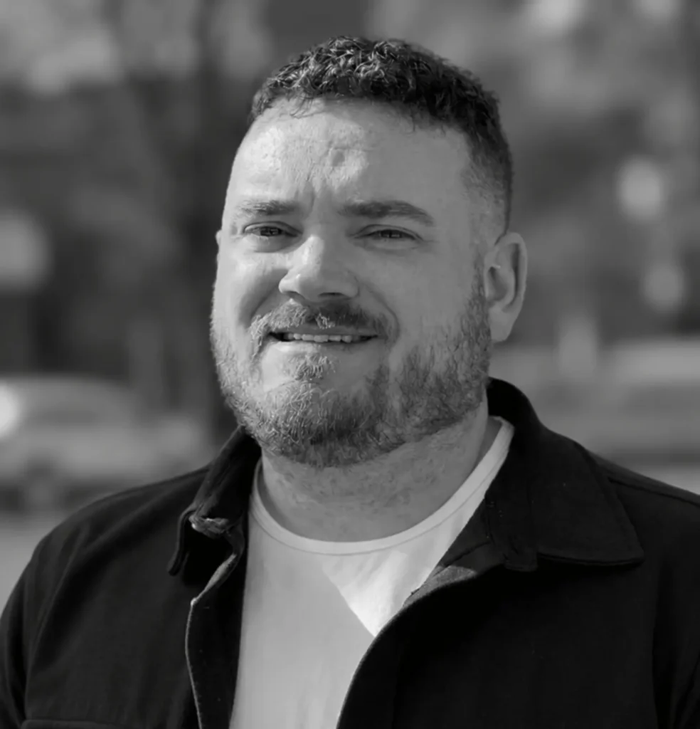 A man with short curly hair and a beard, wearing a light shirt and dark jacket, stands outdoors and smiles at the camera. The image is in black and white.