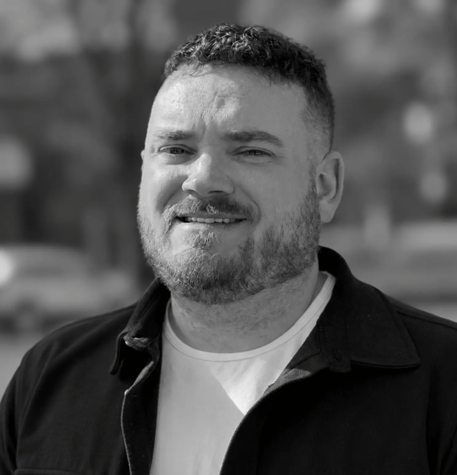 A man with short curly hair and a beard, wearing a light shirt and dark jacket, stands outdoors and smiles at the camera. The image is in black and white.