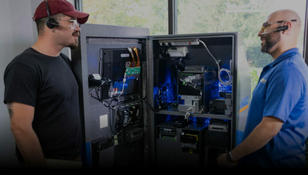 Two men wearing smart glasses and headsets stand next to an open electronics cabinet, examining its internal components in a well-lit room.