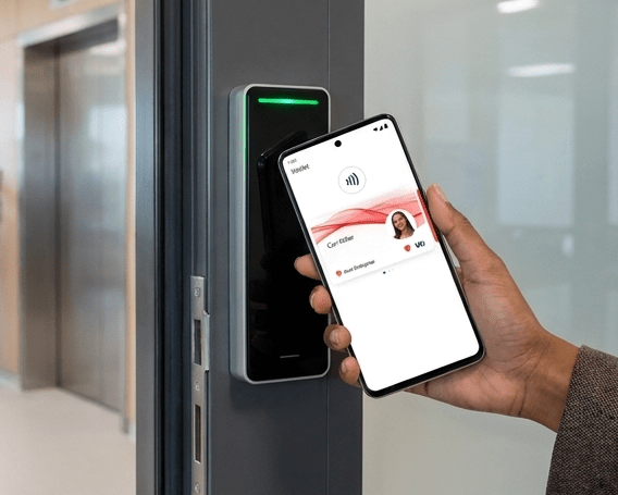 A hand holds a smartphone with a digital employee badge displayed, tapping it to an electronic access control reader near a door.